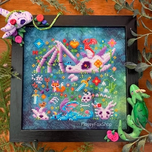 May include: A framed cross-stitch artwork featuring a whimsical dragon surrounded by colorful flowers and other fantasy elements. The dragon is primarily white and purple, set against a blue-green background. The frame is black, and the artwork is embellished with decorative clay figures.