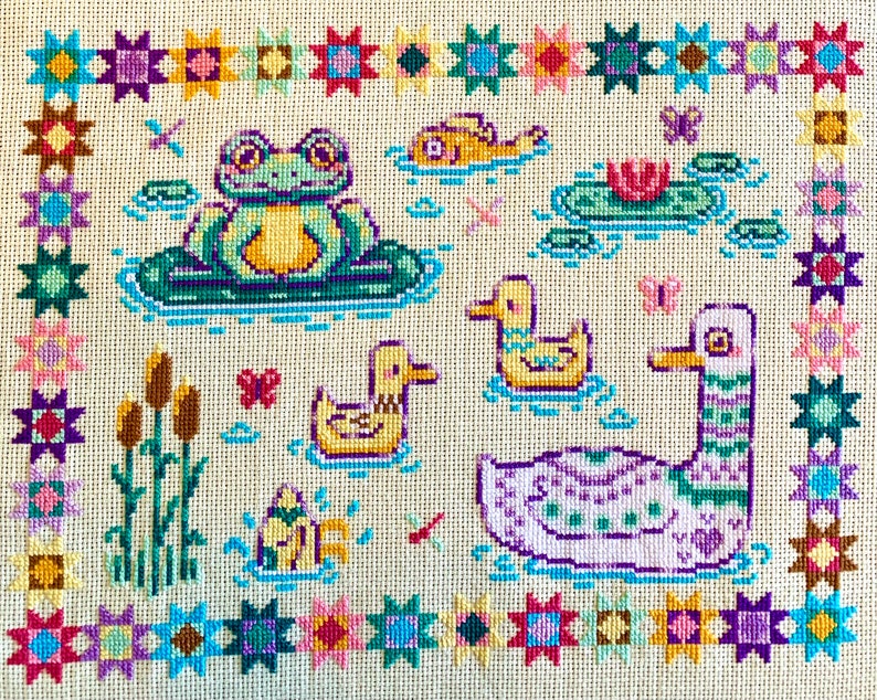 May include: A cross-stitch embroidery design featuring a frog sitting on a lily pad, a duck, a swan, and a variety of flowers and plants. The design is surrounded by a border of colorful stars.