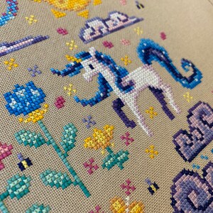 Puede incluir: Bordado de punto de cruz con un unicornio blanco con una crin y cola azules, rodeado de flores amarillas, flores azules y nubes. El sol est&aacute; en la esquina superior izquierda de la imagen.