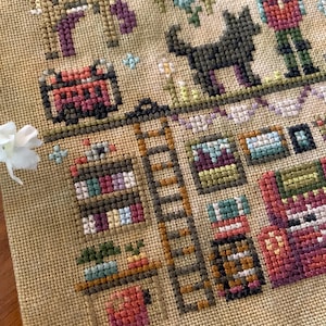 May include: A detailed cross-stitch sampler featuring various figures and objects. The design includes a brown deer, a black dog, a person, a ladder, bookshelves, and a red cart. The color palette consists of muted tones of brown, purple, green, and red.