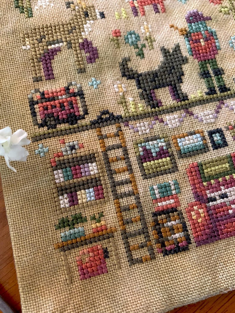 May include: A detailed cross-stitch sampler featuring various figures and objects. The design includes a brown deer, a black dog, a person, a ladder, bookshelves, and a red cart. The color palette consists of muted tones of brown, purple, green, and red.