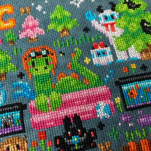 May include: Cross-stitch pattern featuring a green dragon with a pink bed, a black cat, a pink and white snowman, a green tree, and a blue and white creature. The background is a dark blue fabric.