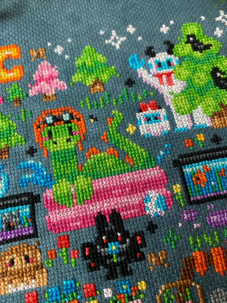 May include: Cross-stitch pattern featuring a green dragon with a pink bed, a black cat, a pink and white snowman, a green tree, and a blue and white creature. The background is a dark blue fabric.