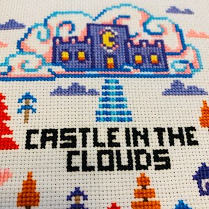 May include: Cross-stitch embroidery featuring a blue castle with a yellow crescent moon, set against a backdrop of pink and blue clouds. The text "CASTLE IN THE CLOUDS" is stitched in black. The design includes trees and houses.