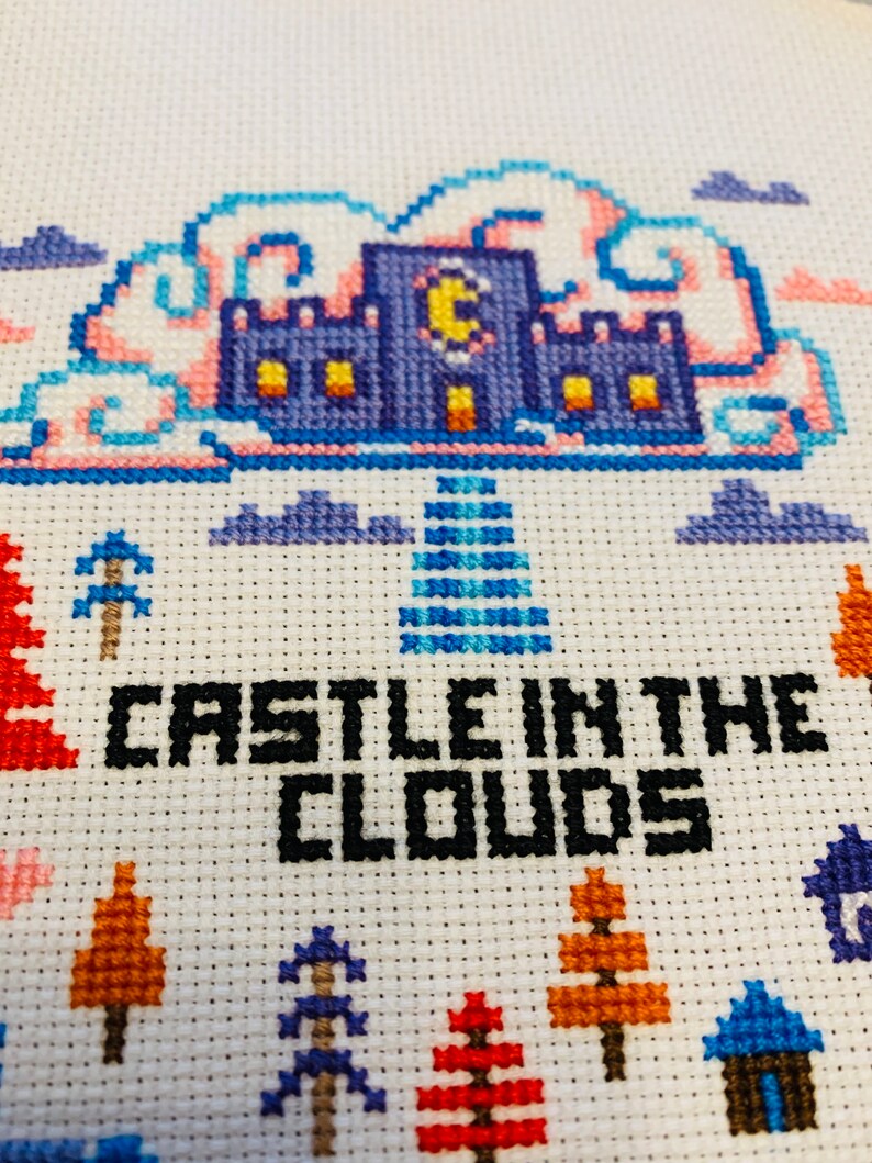 May include: Cross-stitch embroidery featuring a blue castle with a yellow crescent moon, set against a backdrop of pink and blue clouds. The text "CASTLE IN THE CLOUDS" is stitched in black. The design includes trees and houses.