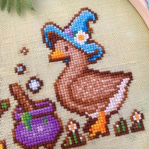 May include: Cross-stitch embroidery of a brown duck wearing a blue witch hat with a white flower. A purple cauldron and small flowers are also stitched. The embroidery is set in a wooden hoop on a light green fabric.