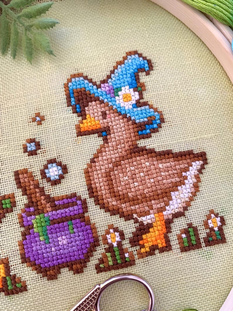 May include: Cross-stitch embroidery of a brown duck wearing a blue witch hat with a white flower. A purple cauldron and small flowers are also stitched. The embroidery is set in a wooden hoop on a light green fabric.