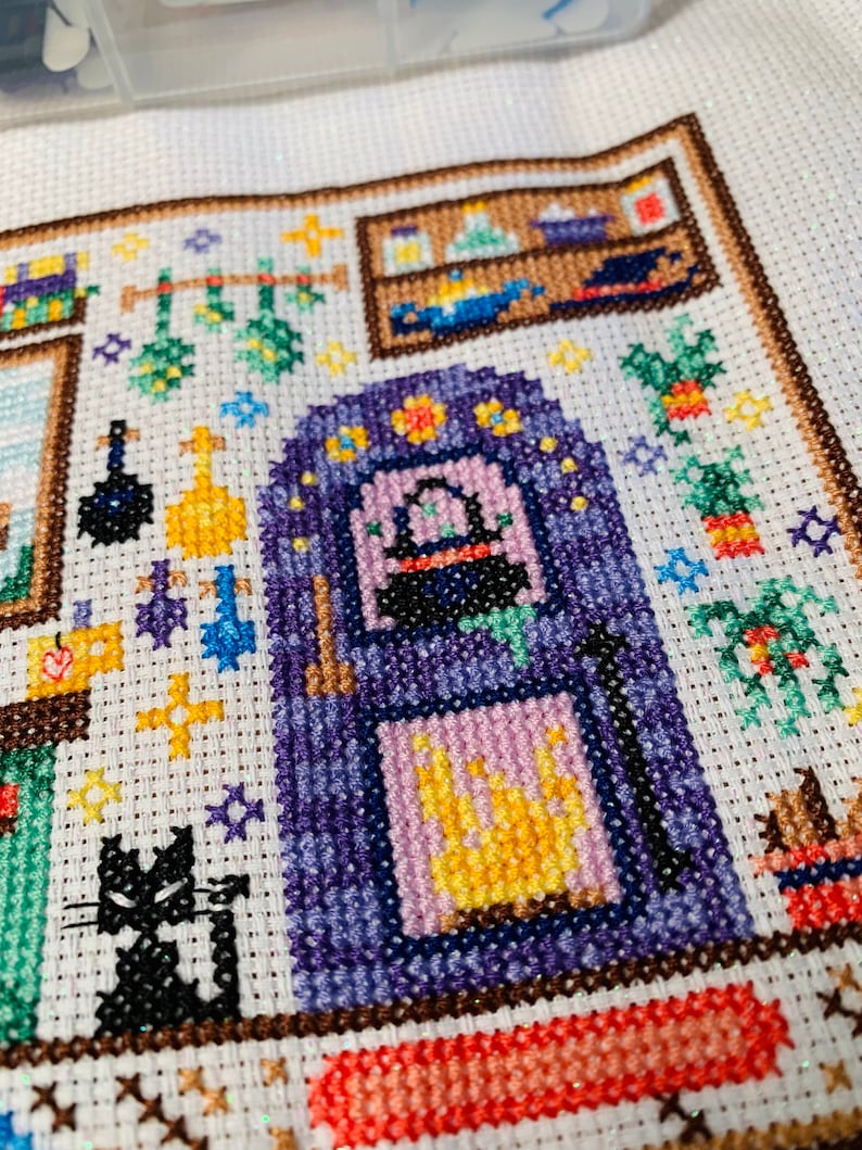 May include: A cross-stitch pattern of a witch's kitchen with a black cat, a cauldron, and a variety of herbs and potions.