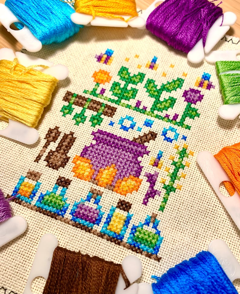 K&ouml;nnte beinhalten: Kreuzstichmuster mit einem violetten Kessel, orangefarbenen K&uuml;rbissen, gr&uuml;nen Pflanzen und blauen und gelben Zaubertrankflaschen. Das Muster ist umgeben von Spulen mit Stickgarn in verschiedenen Farben, darunter violett, orange, gelb, blau und braun.