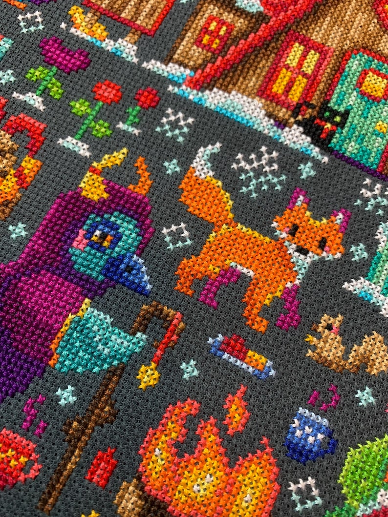 May include: A colorful cross-stitch pattern featuring a fox, a bird, a campfire, and a house. The pattern is on a dark gray background.