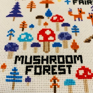 May include: Cross-stitch embroidery featuring a whimsical forest scene. The design includes colorful mushrooms, trees, a deer, and the text "MUSHROOM FOREST". The color palette includes red, blue, orange, and brown on a white background.