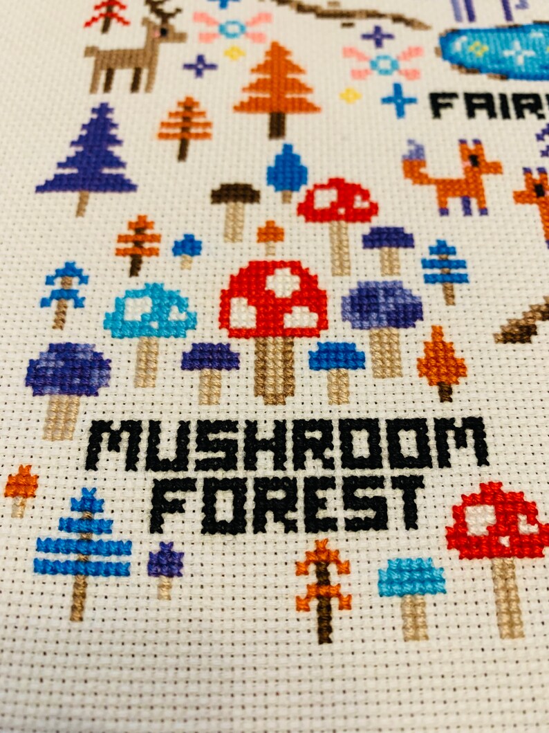 May include: Cross-stitch embroidery featuring a whimsical forest scene. The design includes colorful mushrooms, trees, a deer, and the text "MUSHROOM FOREST". The color palette includes red, blue, orange, and brown on a white background.