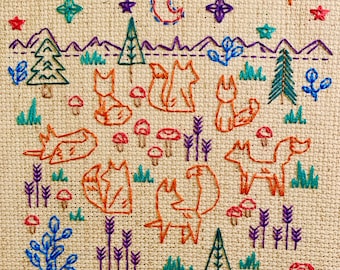 May include: Embroidered design on a beige fabric featuring a moon, stars, trees, and foxes. The foxes are orange and brown with white accents. The design is surrounded by a border of green and blue.