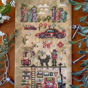 May include: A cross-stitch sampler on tan fabric featuring a whimsical village scene with buildings, cars, animals, and a person. The design includes colorful flowers, a black cat, and a cozy interior with a stove and bookshelves. The text "FlossyFoxShop" is visible.