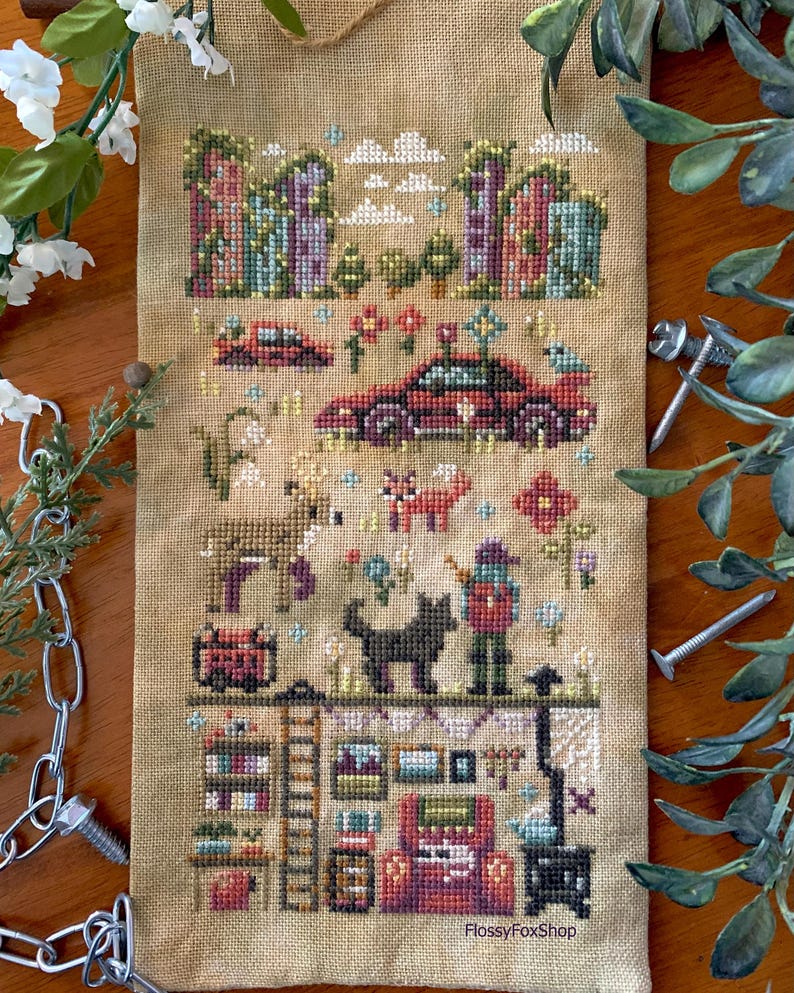 May include: A cross-stitch sampler on tan fabric featuring a whimsical village scene with buildings, cars, animals, and a person. The design includes colorful flowers, a black cat, and a cozy interior with a stove and bookshelves. The text "FlossyFoxShop" is visible.
