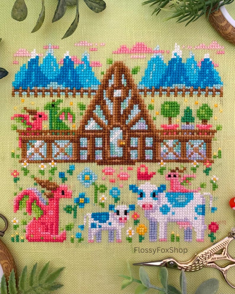 May include: Cross-stitch embroidery featuring a whimsical scene with a brown house, blue mountains, and pink clouds. A pink dragon and a cow with blue spots are in the foreground, surrounded by flowers. The text "FlossyFoxShop" is visible.