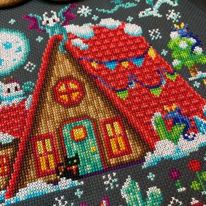 May include: A cross-stitch pattern of a cozy winter cabin with a red roof, a green door, and two windows. The cabin is surrounded by snow and decorated with festive flags. A black cat sits in front of the cabin, and a fox and a cactus are in the foreground.