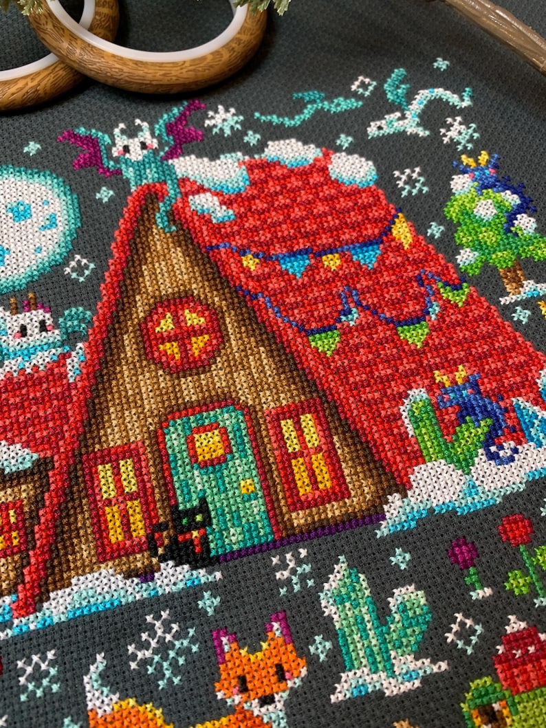May include: A cross-stitch pattern of a cozy winter cabin with a red roof, a green door, and two windows. The cabin is surrounded by snow and decorated with festive flags. A black cat sits in front of the cabin, and a fox and a cactus are in the foreground.