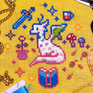 Puede incluir: Un patrón de punto de cruz con un dragón blanco con detalles en rosa y morado, rodeado de estrellas, una espada, una maceta, un hongo y una taza. El dragón está sentado en un cofre azul y rosa.