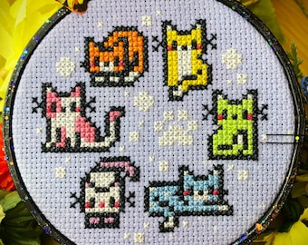 May include: A purple embroidery hoop with a cross-stitch design of six colorful cats on a white background. The cats are in various poses and have different colored fur.