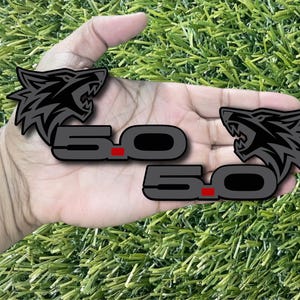 Puede incluir: Dos emblemas negros y grises 5.0 con un diseño de cabeza de lobo. Los emblemas tienen un acento rojo y se sostienen en una mano sobre un fondo verde.