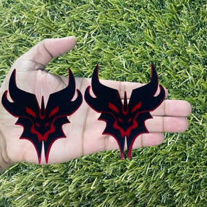 May include: Two black and red decorative emblems with a symmetrical design featuring curved horns and a central face-like shape. The emblems are held in a hand against a backdrop of green artificial grass.
