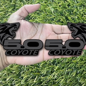May include: Two black and gray 5.0 Coyote decals. Each decal features a wolf head graphic above the text "5.0 Coyote". The decals are displayed on a green, artificial grass surface.