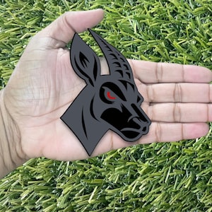 Può includere: Un adesivo piatto fustellato con una grafica stilizzata di una testa di gazzella o antilope grigio scuro di profilo. Il design presenta contorni neri netti, un corno ricurvo e un singolo occhio rosso penetrante, creando un aspetto audace.