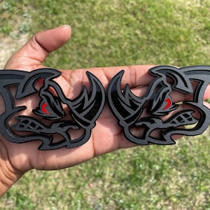 Może przedstawiać: Dwa czarne, szczegółowe, trójwymiarowe emblematy samochodowe z czerwonym okiem i podobnym do nosorożca obliczem.