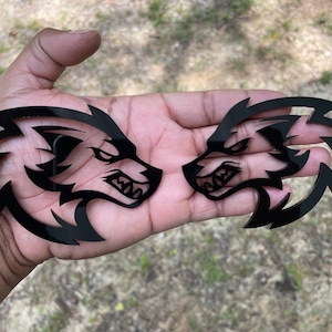 May include: Two black wolf head emblems with a fierce expression. The emblems are mirror images, with sharp lines and a glossy finish. The wolf heads face each other, creating a dynamic and aggressive design. The emblems are likely for vehicle decoration.