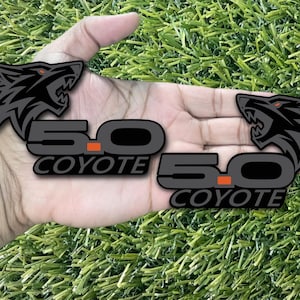 Puede incluir: Dos pegatinas con el emblema 5.0 Coyote en negro y gris con un diseño de cabeza de lobo. Las pegatinas tienen un acento naranja y se muestran sobre una superficie de césped artificial verde.