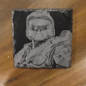 May include: A black slate coaster featuring a grey engraved image of a video game character wearing a helmet. The character is facing forward and holds a gun in their right hand.