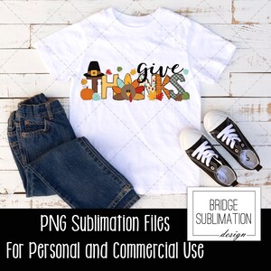 May include: White t-shirt with the words "give thanks" in a colorful Thanksgiving-themed design. The design includes a turkey, a pilgrim hat, and fall leaves. The image also shows a pair of blue jeans and black sneakers.