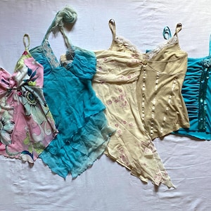 May include: Four sleeveless tops arranged side by side. The first is pink with a large floral print. The second is a turquoise halter top with layered fabric. The third is a beige asymmetrical top with floral details. The fourth is a bright blue tank top.