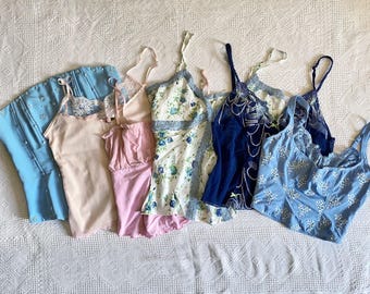 May include: A collection of camisoles and lingerie in various colors, including light blue, pink, and navy. Some pieces feature floral patterns, lace trim, and delicate straps. The garments are arranged on a white, textured surface.