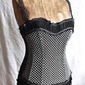 May include: Black corset top with a white diamond pattern, ruffled trim, and thin straps. The top is displayed on a vintage mannequin. The corset features a structured bodice and delicate detailing.