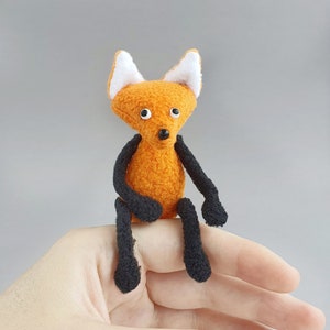 Puede incluir: Un peluche de zorro naranja hecho a mano con brazos y piernas negros y orejas con puntas blancas. El zorro tiene una pequeña nariz negra y está sentado en un dedo. El juguete está hecho de tela suave y texturizada.