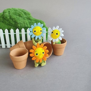 Może przedstawiać: Trzy postaci kwiatów wykonanych na szydełku w doniczkach z terakoty. Kwiaty są żółte, pomarańczowe i białe z zielonymi łodygami i twarzami. Doniczki stoją na szarym tle z zielonym krzewem i białym drewnianym płotem w tle.