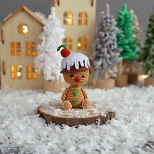 May include: A crocheted gingerbread person doll with a white icing hat and a red candy on top. The doll is sitting on a wooden slice in front of a miniature winter village scene.
