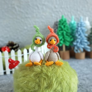 Puede incluir: Dos figuritas de ganso amigurumi de crochet con sombreros verdes y naranjas sentadas sobre una superficie verde peluda. Los gansos están sentados frente a una valla blanca de madera y una fila de árboles en miniatura.