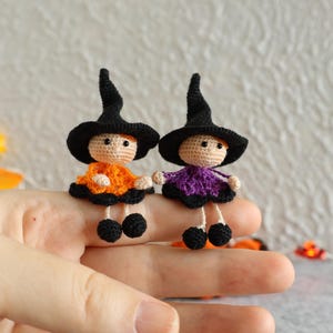 Può includere: Due piccole bambole da strega all'uncinetto fatte a mano. Una bambola indossa un vestito arancione, l'altra un vestito viola, entrambi con dettagli neri. Ogni bambola ha un cappello da strega nero e scarpe nere. Le bambole sono esposte su una mano.