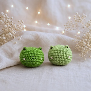 May include: Two handmade green crochet frog toys with small pink accents for the mouth and eyes. The frogs are round and have small ears. They are placed on a neutral-colored fabric with soft lighting and decorative flowers in the background.