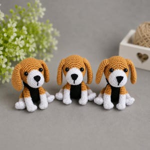 Könnte beinhalten: Drei handgefertigte Häkel-Beagle-Hundefiguren. Jeder Hund hat einen hellbraunen Kopf und Ohren, eine weiße Schnauze und Pfoten sowie einen schwarzen Kragen. Die Hunde sitzen auf einer grauen Oberfläche. Der Hintergrund umfasst Grün und eine Holzkiste.