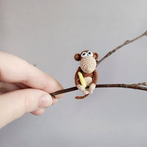 Peut inclure: Petite figurine de singe crocheté, marron et beige, tenant une banane jaune. Le singe est perché sur une brindille brune sur fond neutre. La figurine mesure environ 5 cm de haut.