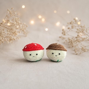 Puede incluir: Dos figuras de setas de ganchillo con caras sonrientes. Una tiene una gorra roja con lunares blancos, la otra una gorra marrón. Ambas tienen cuerpos blanquecinos y bases verdes. El fondo es de un color suave y neutro con luces borrosas.