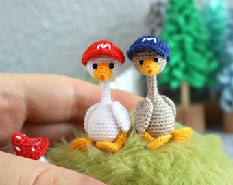 Può includere: Due piccole statuette di oche all'uncinetto, una bianca e una marrone chiaro, che indossano ciascuna un minuscolo berretto con una lettera M bianca ricamata sul davanti. L'oca bianca indossa un berretto rosso e quella marrone un berretto blu.