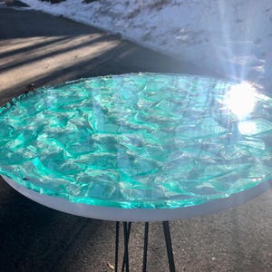 May include: Round side table with a turquoise resin top and white rim. The resin top has a textured appearance. The table has black hairpin legs. The table is outdoors on a sunny day.