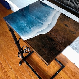 May include: A dark wood end table with a black metal frame. The tabletop features a resin design of a blue ocean wave crashing on a white sandy beach.