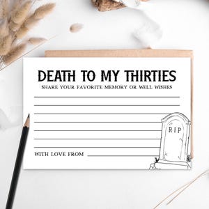 May include: A white card with the text "DEATH TO MY THIRTIES" and "SHARE YOUR FAVORITE MEMORY OR WELL WISHES". The card has lines for writing and a tombstone illustration. A pencil and dried plants are in the background.