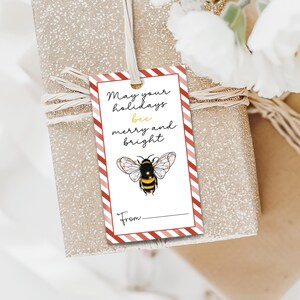 以下が含まれることがあります： 赤と白のストライプのボーダーと「May your holidays bee merry and bright」のテキストが入ったギフトタグ。テキストの下には、ミツバチの詳細なイラストが描かれています。タグは白いリボンで包まれたギフトに結ばれています。